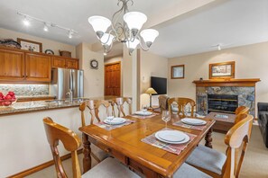 A dining area with a wooden table set for four, surrounded by wooden chairs. A stone fireplace and a television are visible in the background. - Popular Ski-in, Ski-out Location, Shared Hot tub (Whistler)