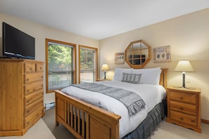 A bedroom with a wooden bed, a TV on a dresser, two bedside tables with lamps, a dresser, and a window with blinds.