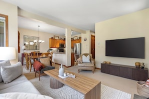 A living room with a sofa, coffee table, and TV. There is a kitchen in the background with wooden cabinets and appliances. - Popular Ski-in, Ski-out Location, Shared Hot tub (Whistler)
