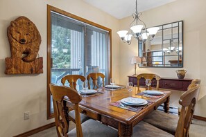 Comedor con mesa de madera dispuesta para cuatro, sillas, lámpara de araña y un espejo grande sobre la mesa.