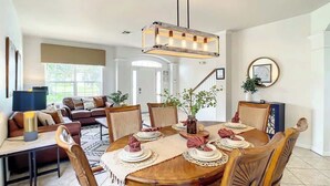 A dining area with a wooden table set for six, surrounded by wooden chairs. There is a chandelier above the table, and a plant is placed on the table.