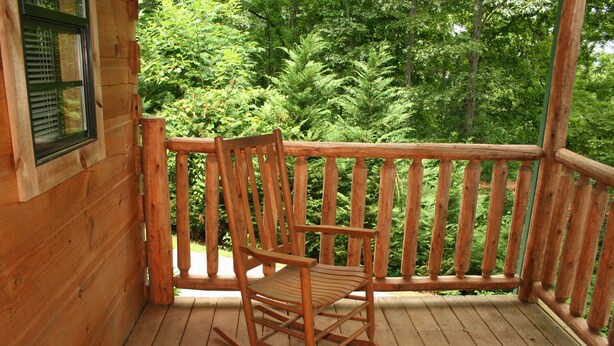A wooden rocking chair on a wooden deck with a railing, surrounded by dense green foliage. - Bear Cabin - 2 Bedroom/1 Bath Vacation Rental (Franklin)