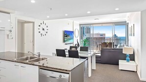 A modern kitchen with a dishwasher, a dining area with a table and chairs, a sofa, and a large window offering a city view. - Chevron Renaissance Ocean View (Surfers Paradise)