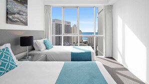 A hotel room with a bed, a nightstand, a lamp, a painting, and a large window with a view of buildings and the sea. - Chevron Renaissance Ocean View (Surfers Paradise)