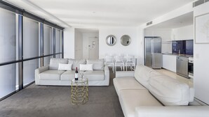 A modern living room with a white sofa, a coffee table with a bottle and glasses, and a kitchen area with white cabinets and appliances. - Circle on Cavill Ocean View (Surfers Paradise)