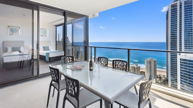 A balcony with a glass table, chairs, and a bed with a blue blanket. - Circle on Cavill Ocean View (Surfers Paradise)