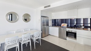 A modern kitchen with white cabinets, a stainless steel refrigerator, an oven, and a dining area with white chairs and a table. - Circle on Cavill Ocean View (Surfers Paradise)