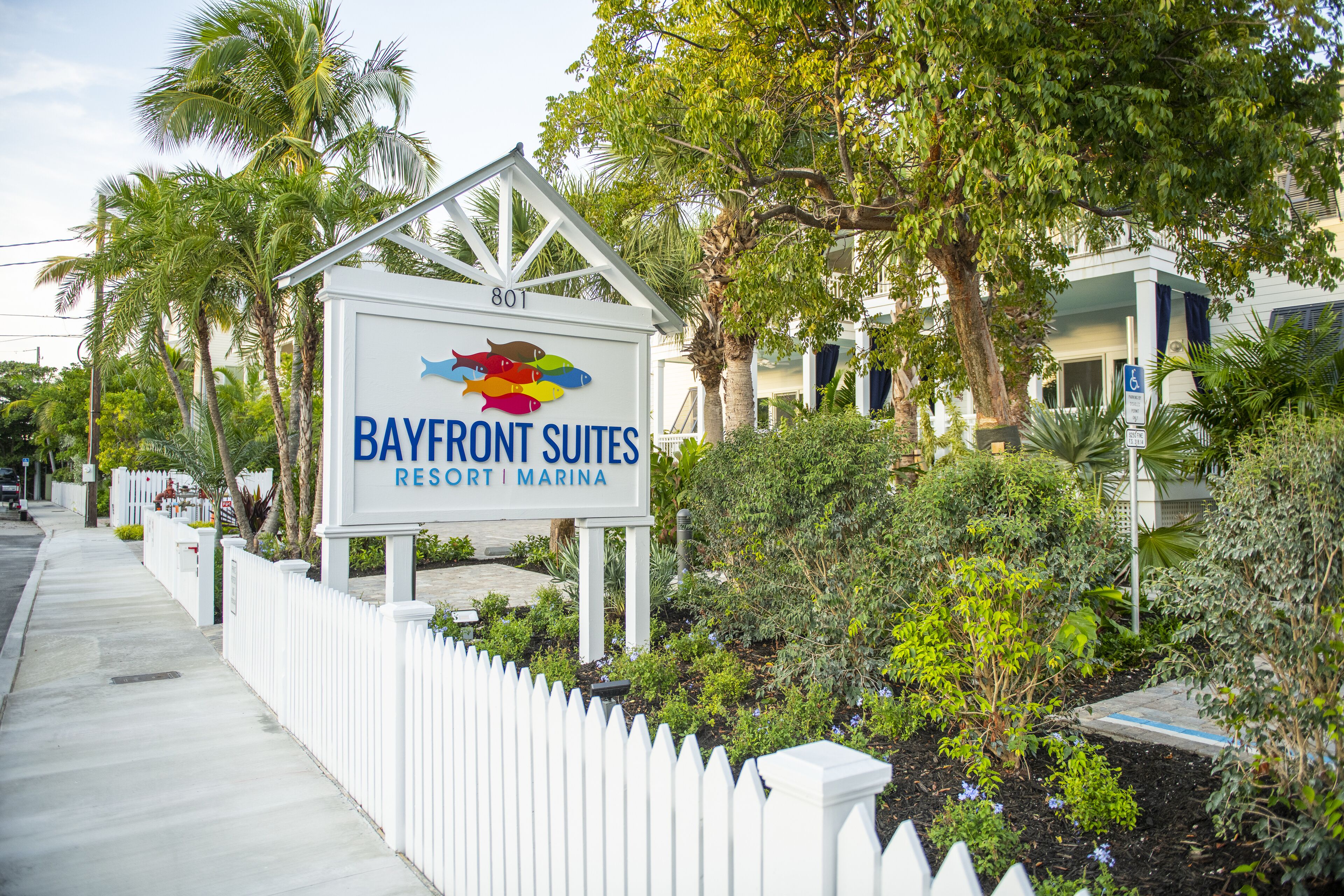A sign for Bayfront Suites Resort & Marina with a white picket fence and lush greenery.