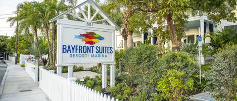 A sign for Bayfront Suites Resort & Marina with a white picket fence and lush greenery.