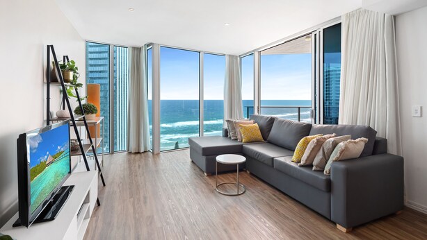A modern living room with a grey sofa, a flat-screen TV, and a view of the ocean through large windows. - Residences H with ocean view (Surfers Paradise)