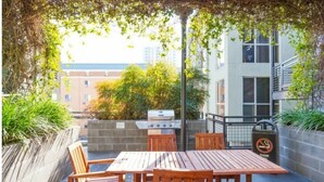 Een terras buiten met een houten tafel en stoelen, een barbecue en een vuilnisbak.