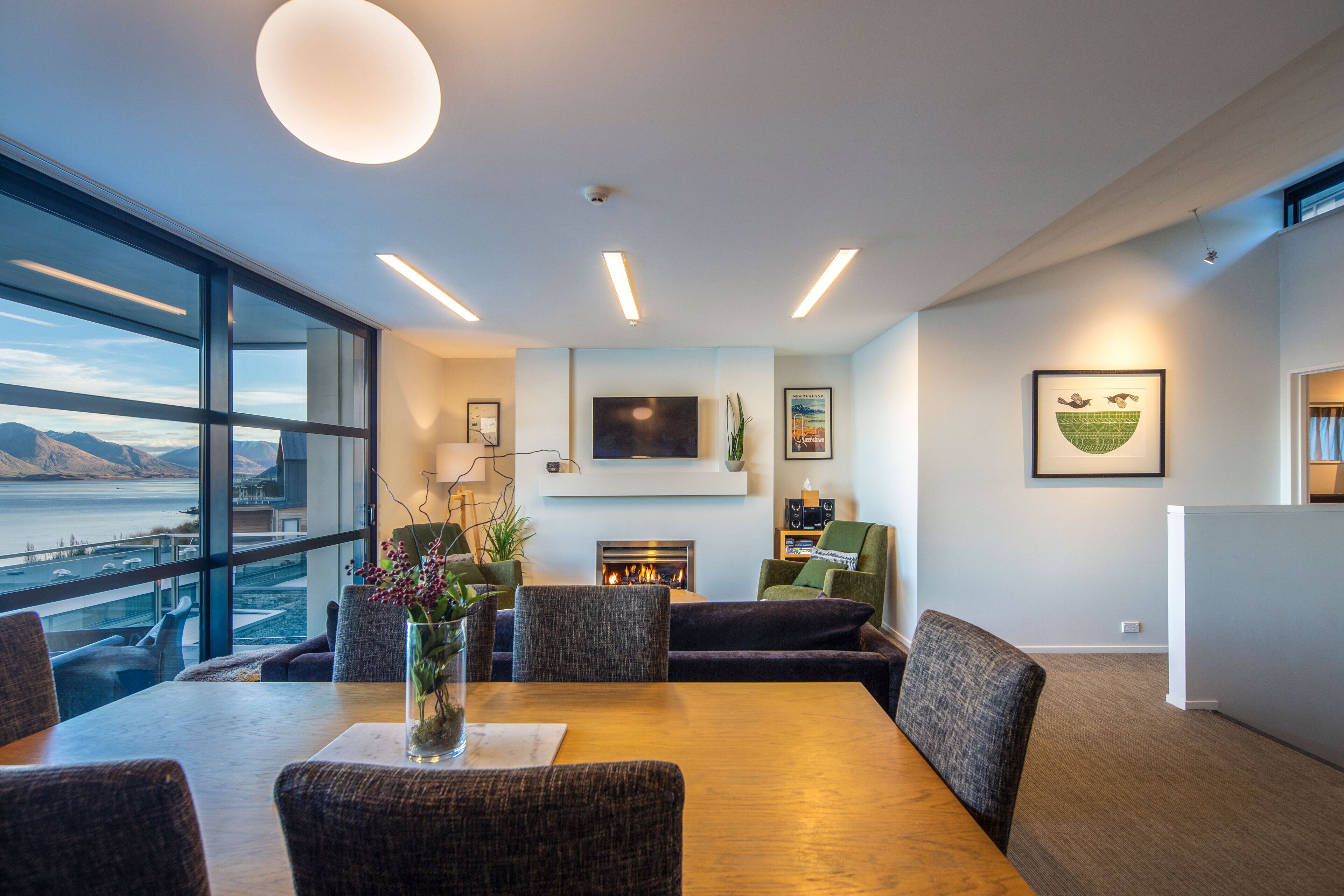 A modern living room with a fireplace, comfortable seating, a large window overlooking a body of water, and a flat-screen TV mounted on the wall.