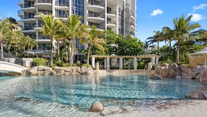 A clear pool with rocks, palm trees, and a building with balconies in the background.
