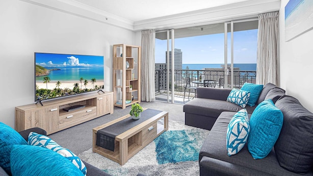 A modern living room with a large flat-screen TV, a wooden entertainment unit, a grey sofa with blue cushions, and a view of the ocean through large sliding doors.