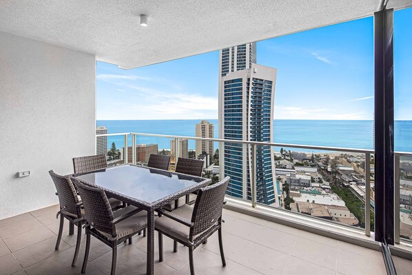 A balcony with a dining table and chairs, overlooking a cityscape and the ocean.