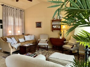Ein Wohnzimmer mit einem beigen Sofa, einem Holztisch, einem Beistelltisch mit Lampe, einem Sessel und einem kleinen Bücherregal.