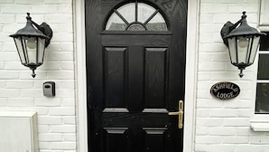 Una puerta negra con un ventanal enmarcado en blanco, flanqueada por dos faroles fijados en la pared. Una placa a la derecha dice 'Ashfield Lodge'.