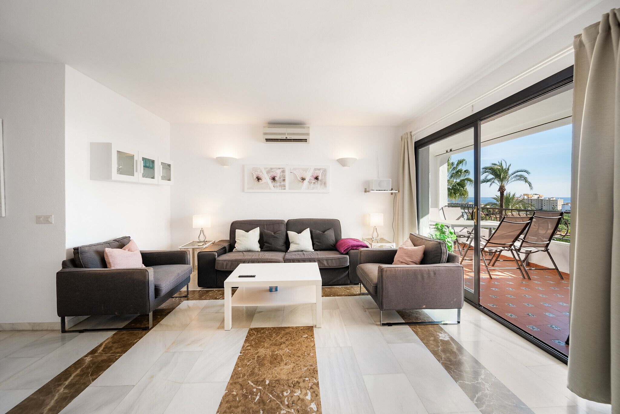 A modern living room with a sofa set, a coffee table, and a large sliding glass door leading to a balcony with outdoor furniture.