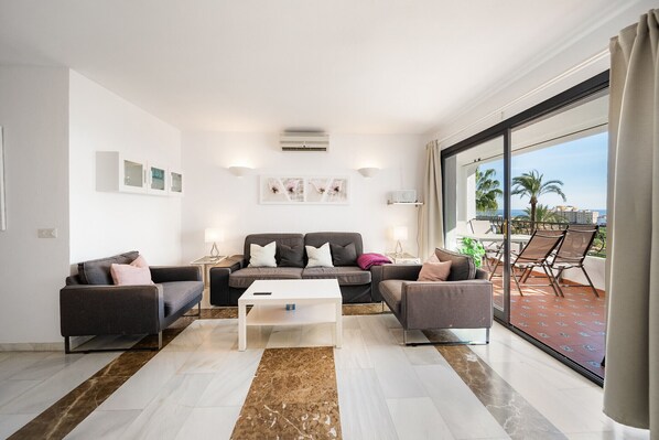 A modern living room with a sofa set, a coffee table, and a large sliding glass door leading to a balcony with outdoor furniture.