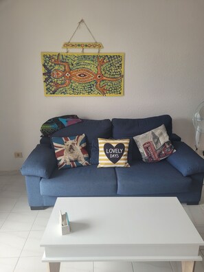 Un sofá azul con varios cojines decorativos, una mesa de centro blanca y un cuadro abstracto colorido en la pared.