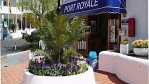 Un jardinière circulaire ornée de fleurs, une marquise bleue avec l’inscription « PORT ROYALE » et un mur blanc.