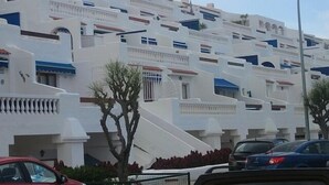 Un immeuble à plusieurs étages aux façades blanches et aux touches de bleu, doté de balcons et d’un espace de stationnement où sont garées des voitures.