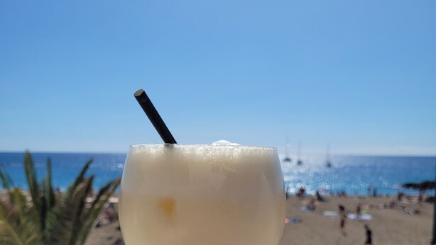 Un verre à cocktail avec une paille, posé sur un balcon donnant sur une plage.