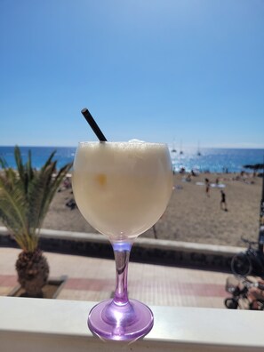 Ein Cocktailglas mit Trinkhalm auf einem Balkon mit Blick auf einen Strand.