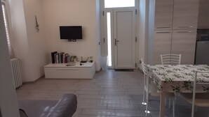 A modern living room with a sofa, a dining table with chairs, a TV mounted on the wall, and a kitchen area with cabinets. - La CasaLina - tourist rental (Omegna)