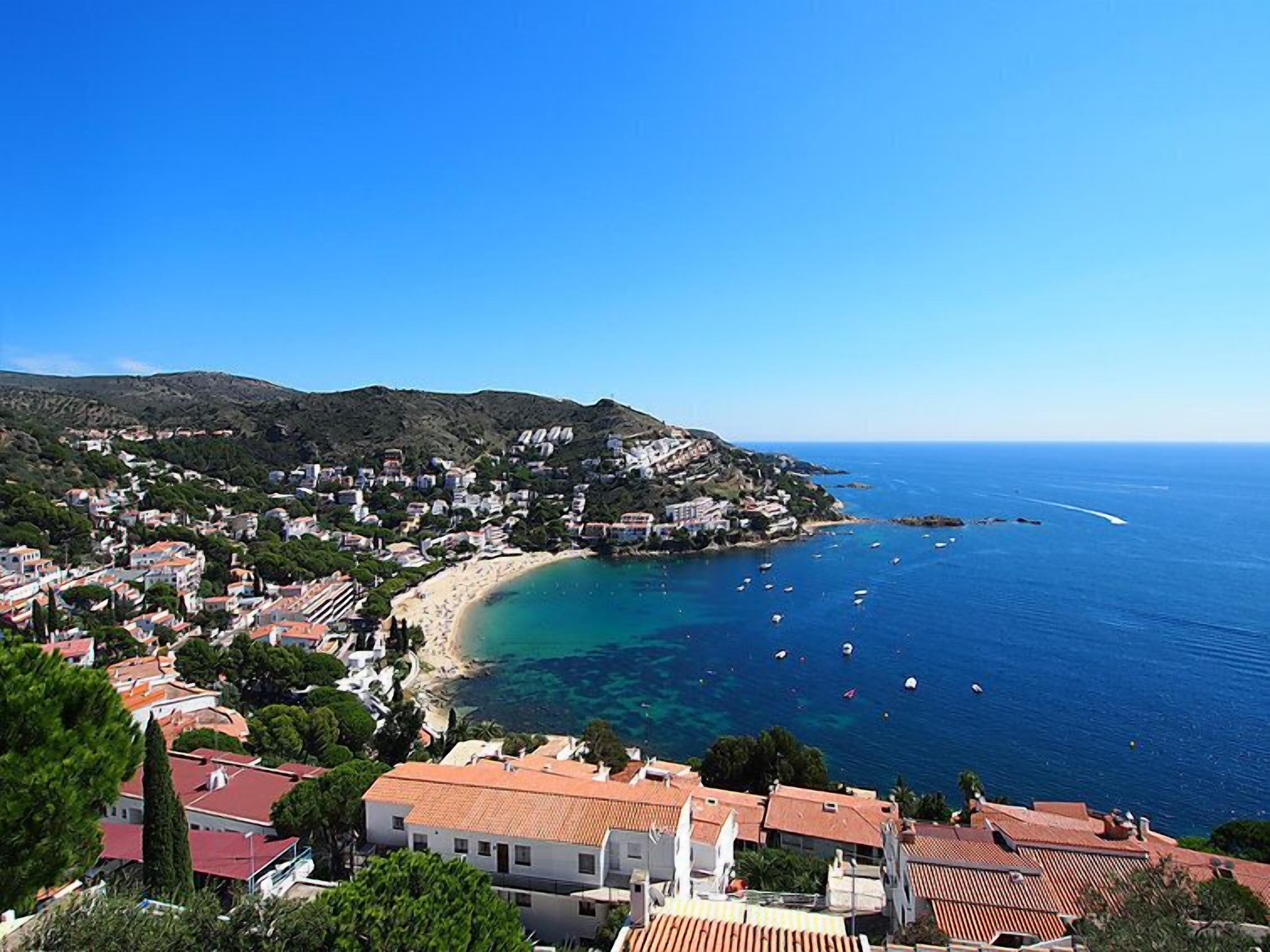 A coastal town with terracotta-roofed buildings, a sandy beach, and clear blue waters with boats.