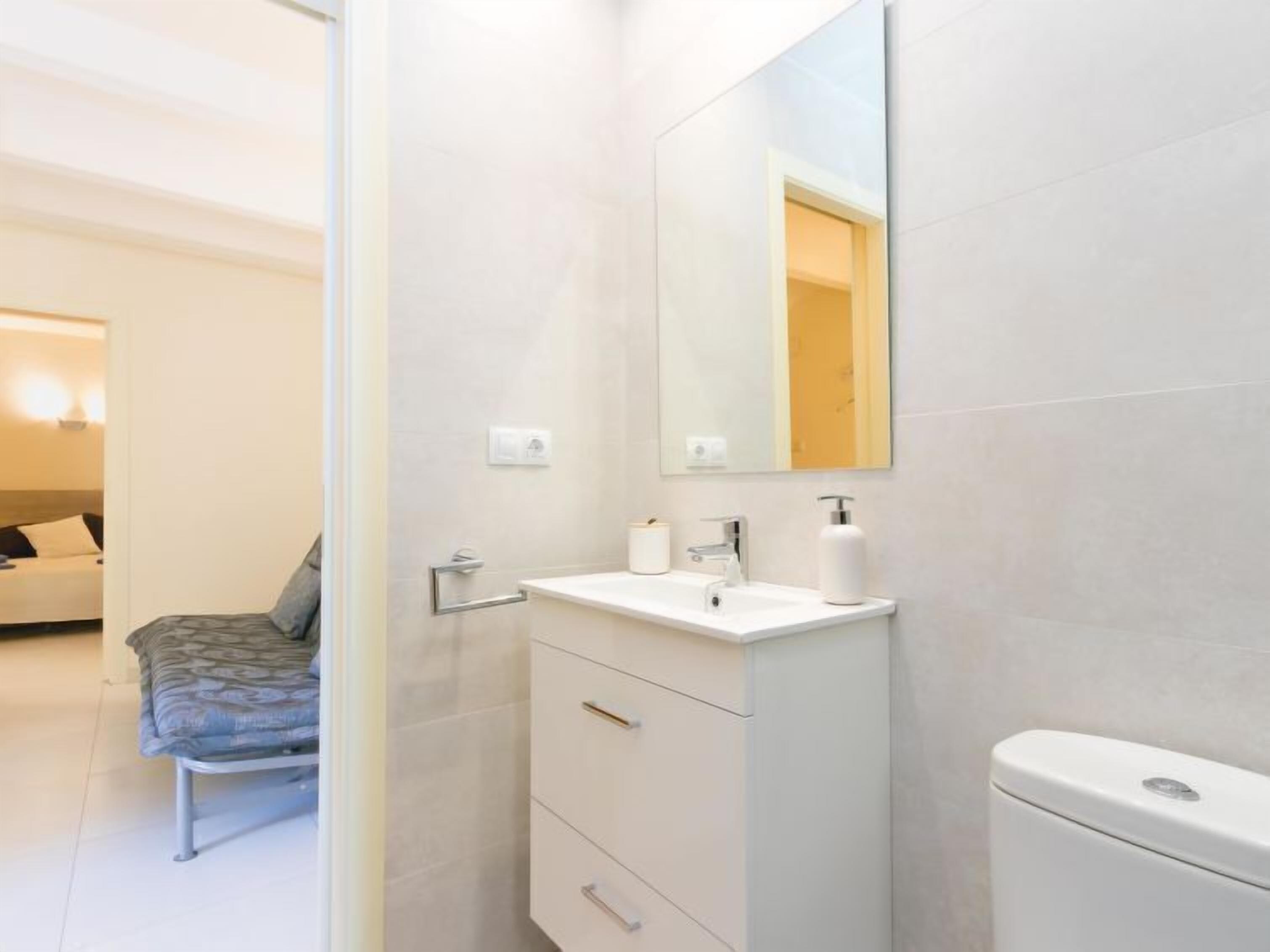 A modern bathroom with a white sink, mirror, and toilet. There is a bench with a patterned cushion in the adjacent room.