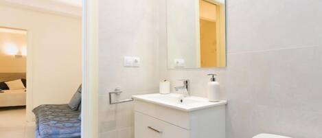 A modern bathroom with a white sink, mirror, and toilet. There is a bench with a patterned cushion in the adjacent room.