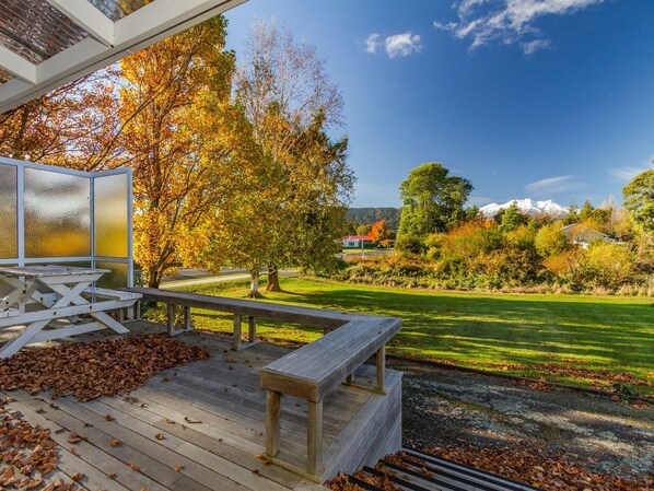 Eine Holzterrasse mit Tisch und Bänken, die auf eine grüne Fläche mit Bäumen und einen entfernten Berg blickt.