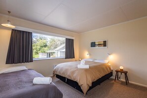 A bedroom with two beds, a window with curtains, a bedside table with a lamp, and a wall-mounted mirror.