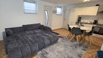 Ett kompakt vardagsrum med en grå soffa, ett litet matbord med stolar, en köksdel med skåp och vitvaror samt ett fönster med gardin.