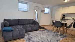 Een kleine woonruimte met een grijze bank, een eettafel met stoelen, een keukenhoek en een deur met kapstok.