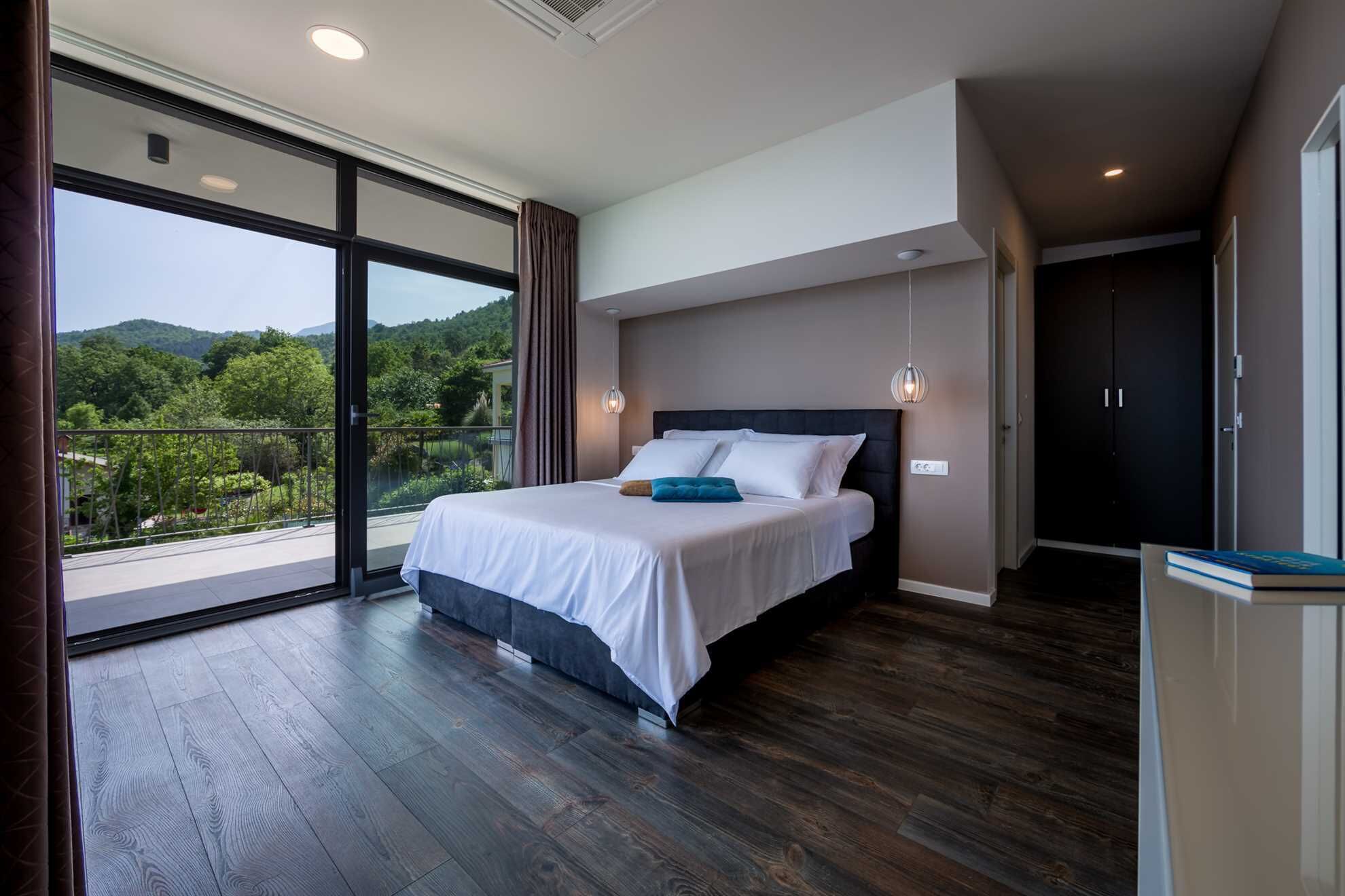 A modern bedroom with a large bed, a balcony with a view of greenery, and a dark wooden floor.
