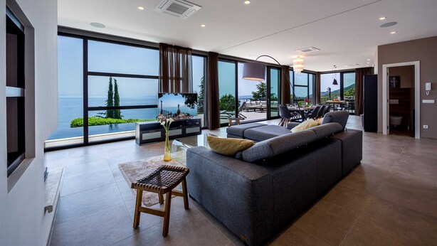 Uma sala de estar ampla com um sofá grande, uma mesa de centro e vista para o mar através de grandes janelas.