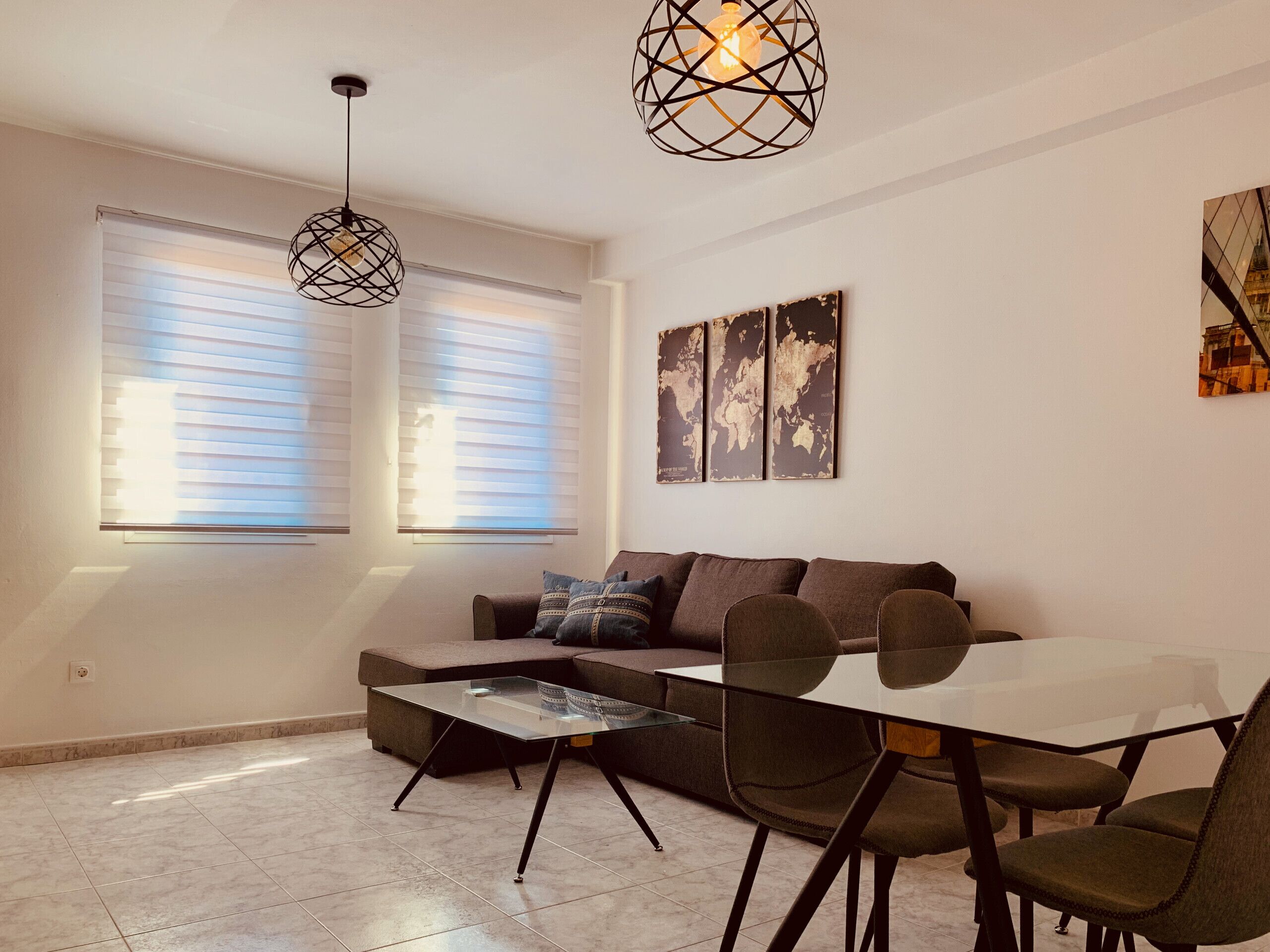 A modern living room with a glass coffee table, a sofa, and a dining table with chairs. There are two large windows with blinds, a world map on the wall, and a hanging light fixture.