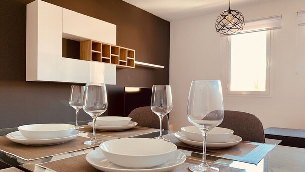 A dining area with a glass table set for four, surrounded by chairs. There is a modern kitchen with white cabinets and a built-in shelf. - A&F Comfort Modern House (Santa Lucía de Tirajana)