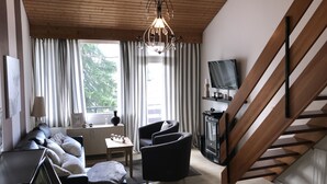 Un salon moderne avec un plafond en bois, un canapé en cuir noir, un fauteuil noir, une petite table et un téléviseur posé sur une étagère en bois.