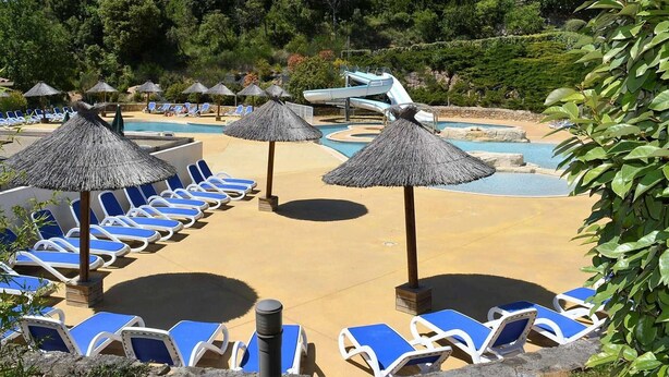 Un área de piscina en un resort, con sillones reclinables y sombrillas de paja.