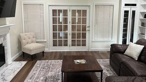 Un salón con chimenea, un sillón beige, un sofá de cuero marrón, una mesa de centro y un ventanal con persianas.