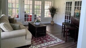 Un salón amplio con un sofá color beige, una mesa de centro y una zona de comedor con una mesa y sillas. Hay ventanas grandes con múltiples paneles y una planta en maceta.