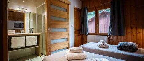 A bedroom with a bed, a window with curtains, a wooden wardrobe, and a bathroom with a glass shower.