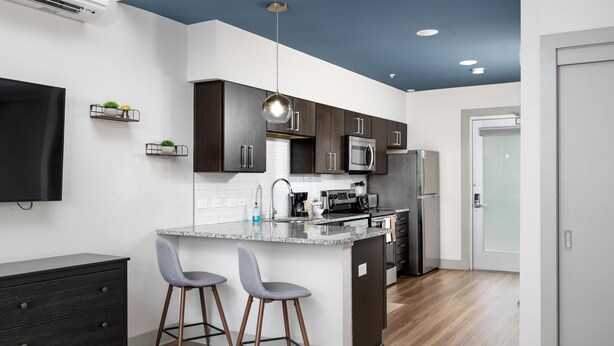 A modern kitchen with dark cabinets, a granite countertop, and stainless steel appliances. There are two bar stools and a small island with a sink. - Studio King Suite with Kitchen | 300 (Asheville)