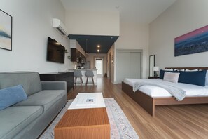 Una habitación de hotel moderna con un sofá gris, una mesa de centro de madera, una cama con ropa de cama azul y blanca, y una zona de cocina al fondo.