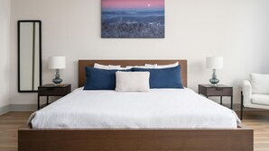 A bedroom with a large bed, two bedside tables with lamps, a nightstand, a chair, and a wall-mounted painting of a landscape. - Studio King Suite with Kitchen | 300 (Asheville)