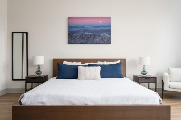 Un dormitorio con una cama grande, dos mesitas de noche con lámparas, una mesita auxiliar, una silla y un cuadro colgado en la pared con un paisaje.