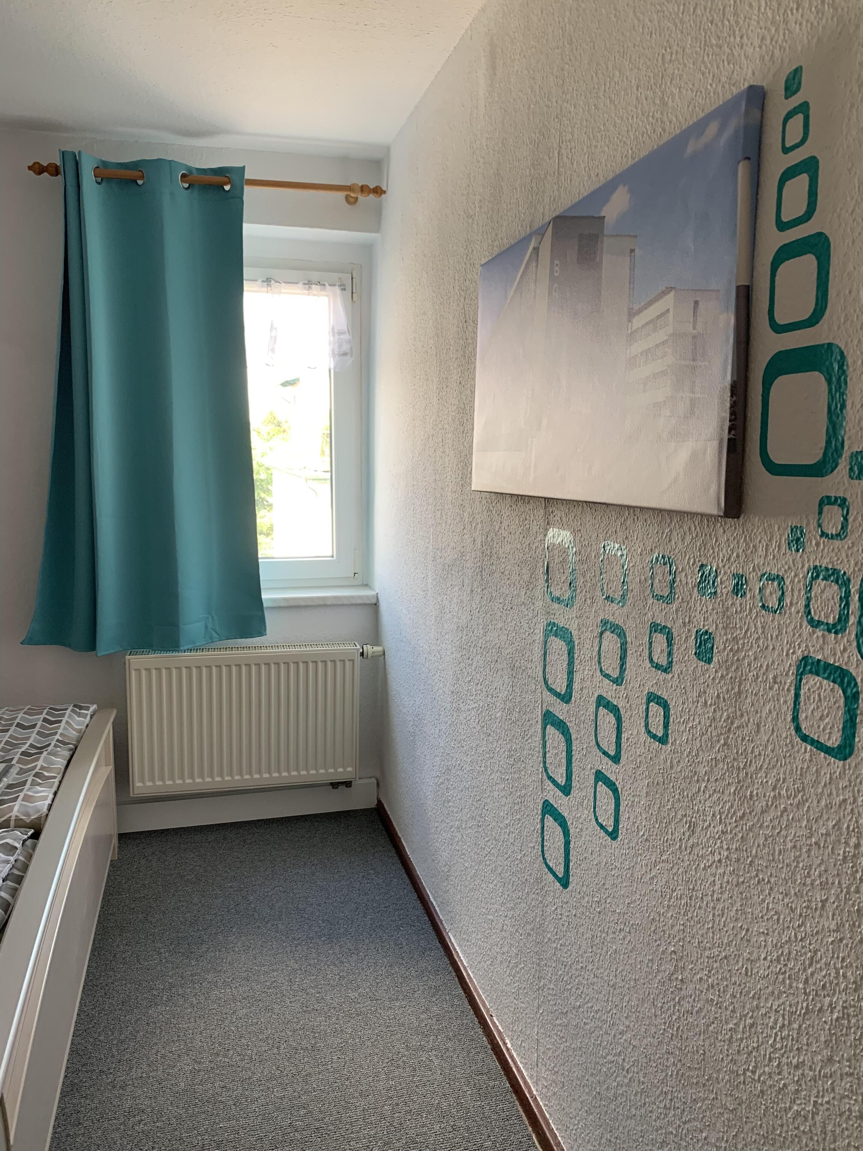 Un dormitorio con cama, ventana con cortinas, radiador y pared con diseño geométrico.